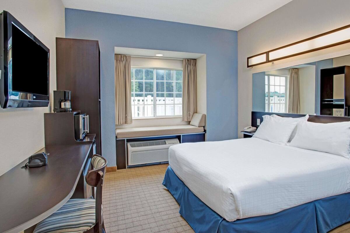 A hotel room with a bed, a desk with a TV and coffee maker, a window with curtains, and a mirror.