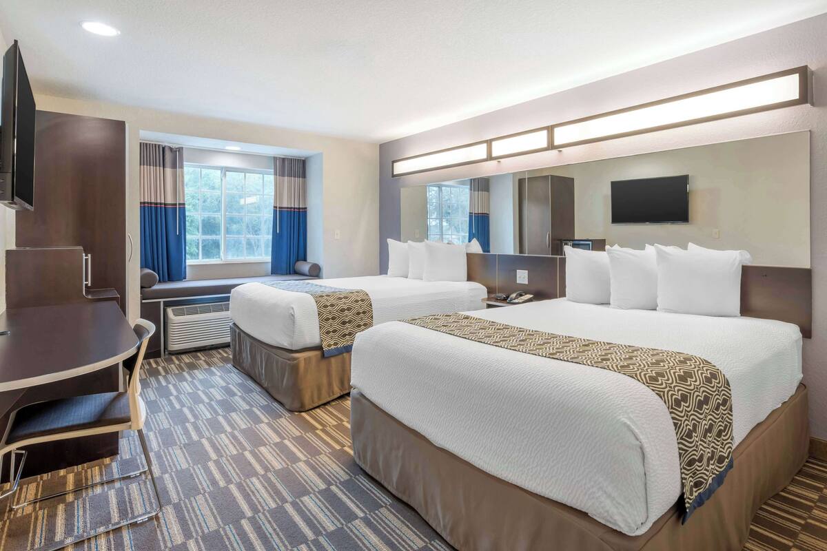 A hotel room with two beds, a desk, a chair, a TV, and a large window with curtains.