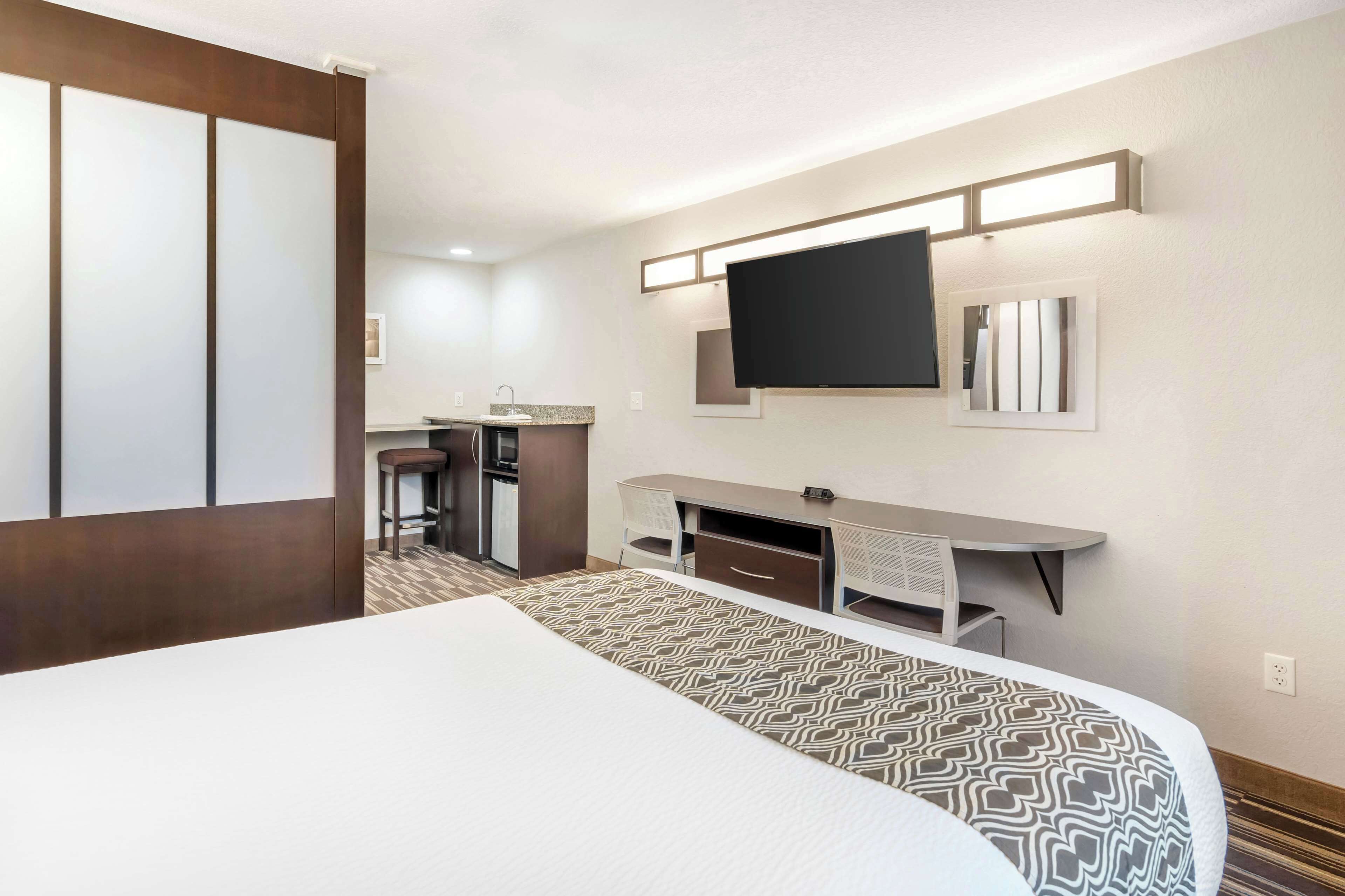 Habitación de hotel con cama, escritorio con silla, televisor montado en la pared y una pequeña zona de kitchenette.