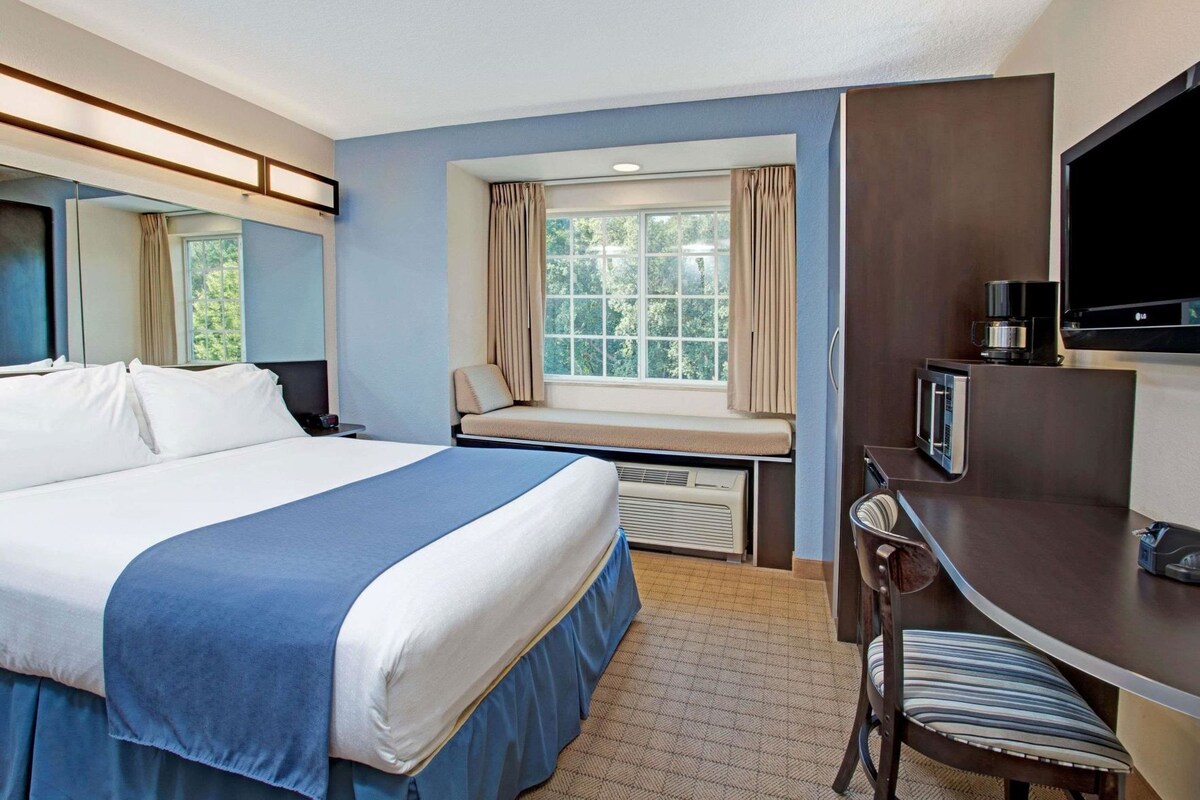 A hotel room with a large bed, a desk with a TV, a window with curtains, and an air conditioning unit.