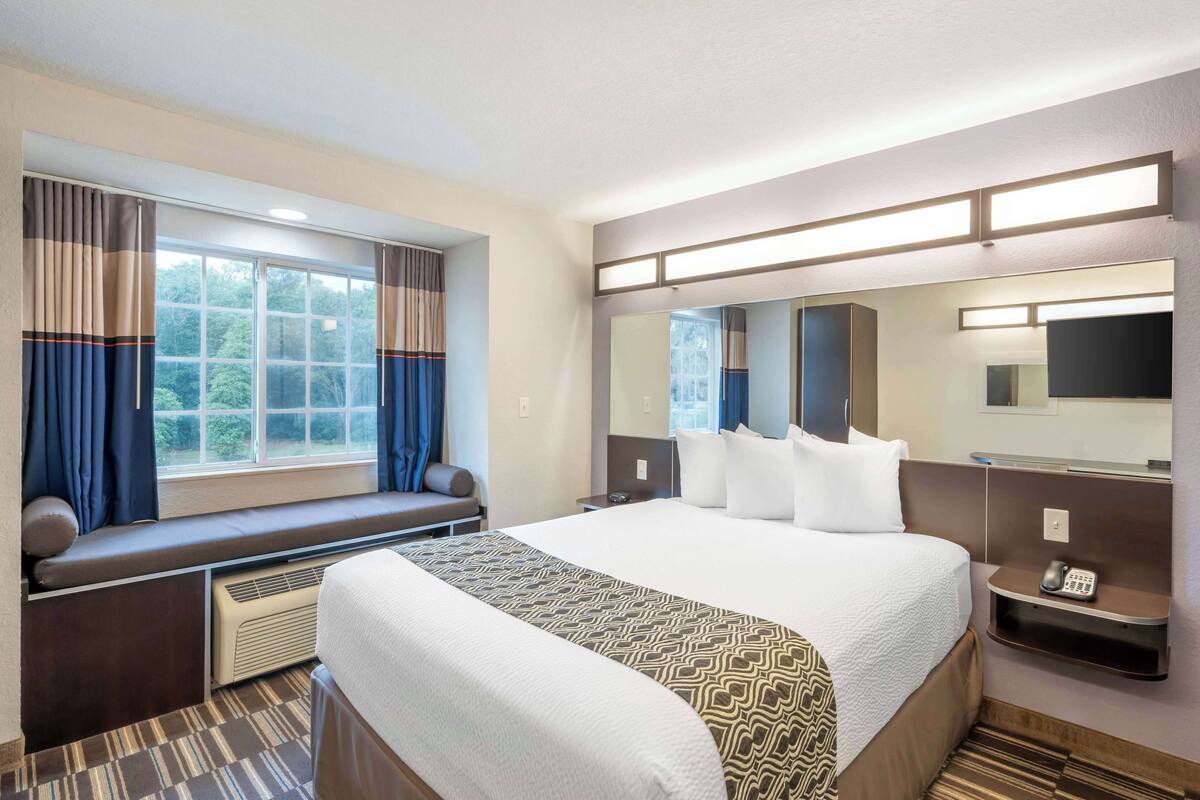 A hotel room with a large bed, a built-in bench with a seat cushion, a large mirror, a flat-screen TV, and a telephone on a bedside table.