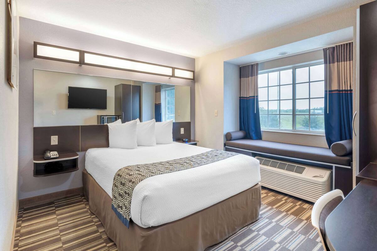 A hotel room with a large bed, a TV mounted on the wall, a window with curtains, a bench by the window, and a desk with a chair.