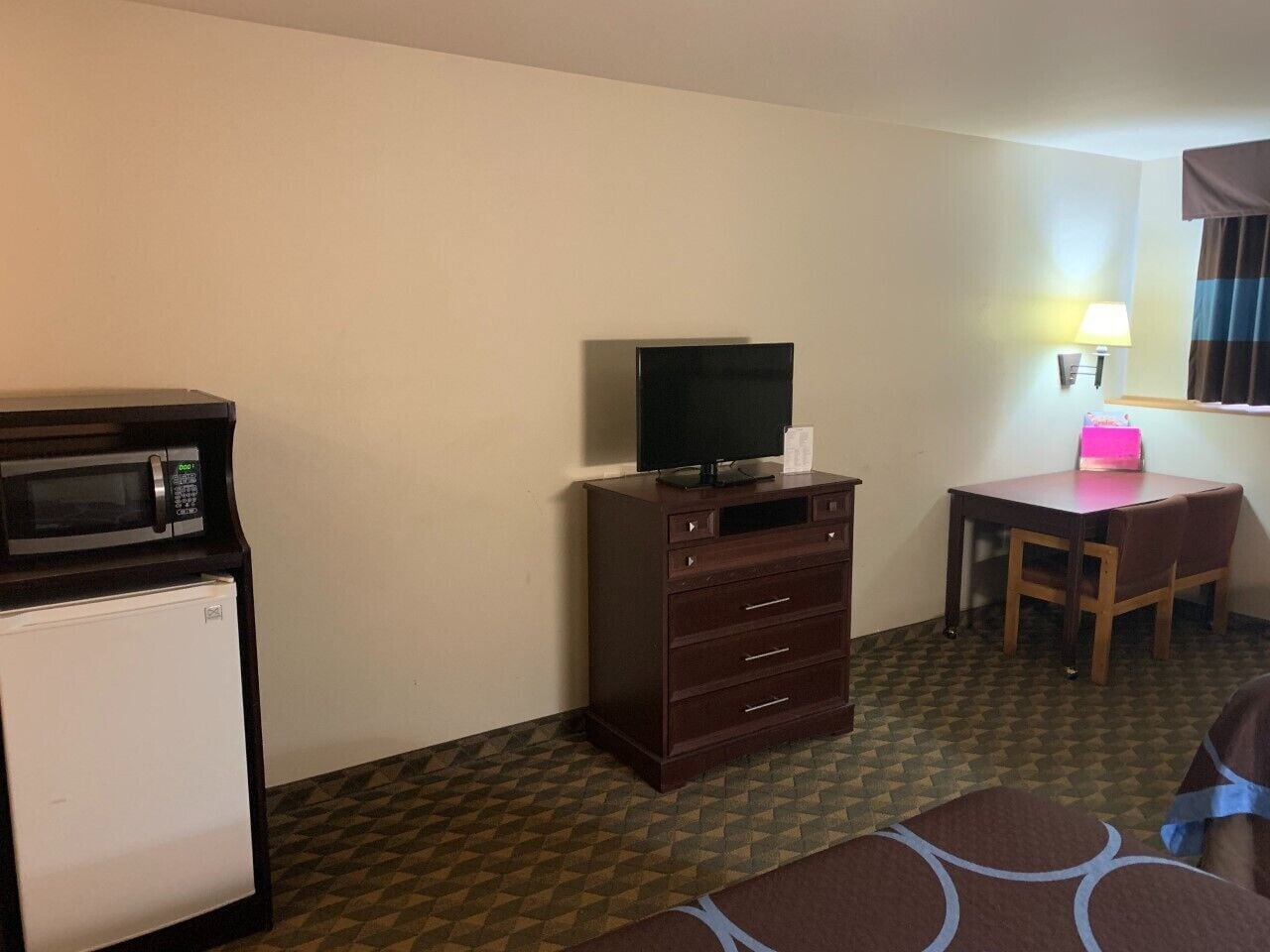 A compact hotel room with a microwave, a small refrigerator, a wooden dresser with a television on top, a desk with a chair, and a lamp.