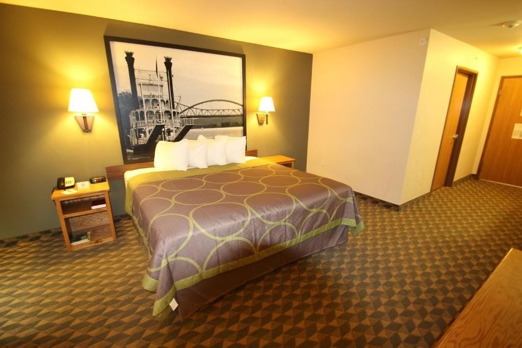 A hotel room with a bed, two bedside lamps, a nightstand, a framed picture on the wall, and a patterned carpet.