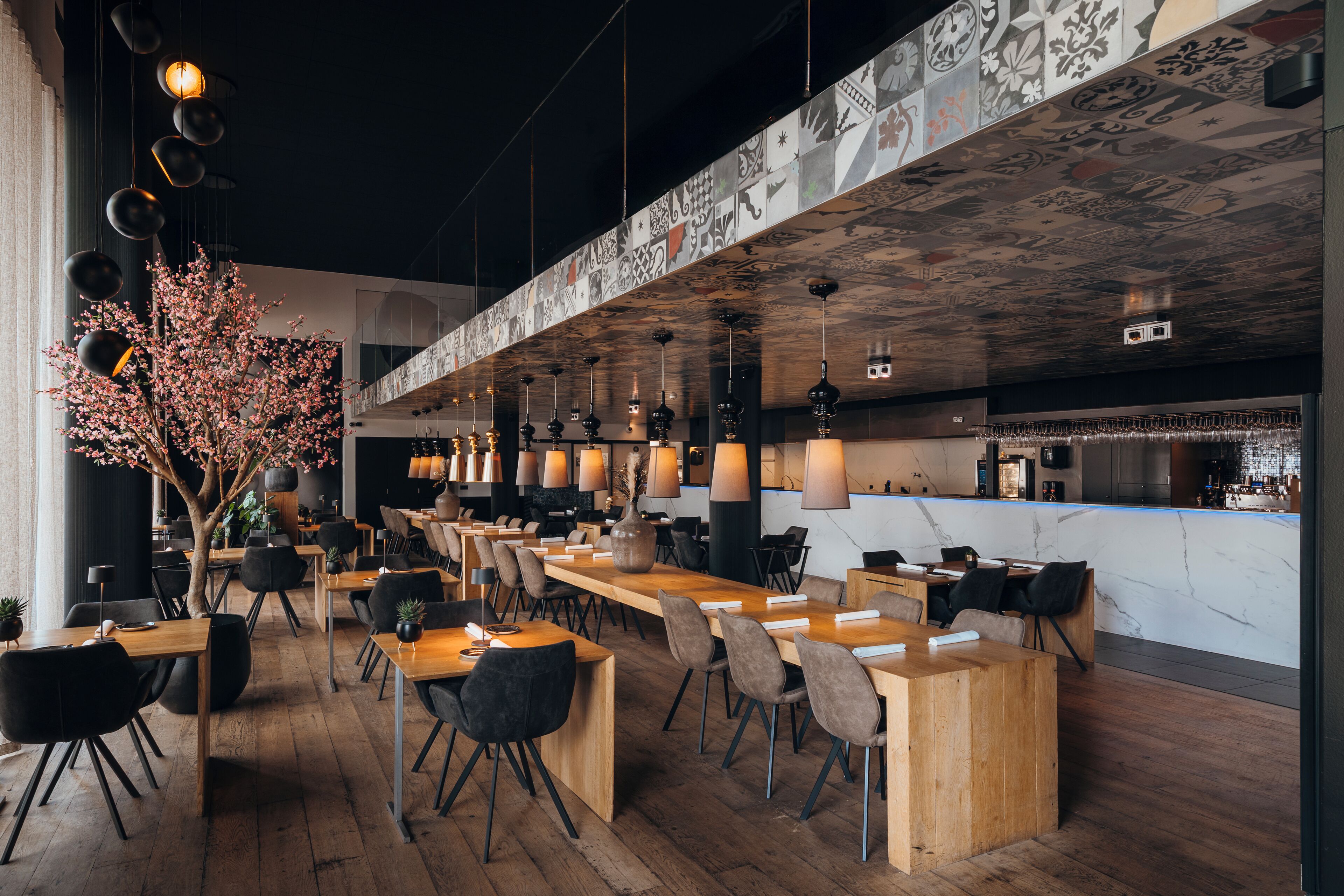 Een modern restaurant met houten tafels en stoelen, hangende pendantlampen en een decoratief plafond.