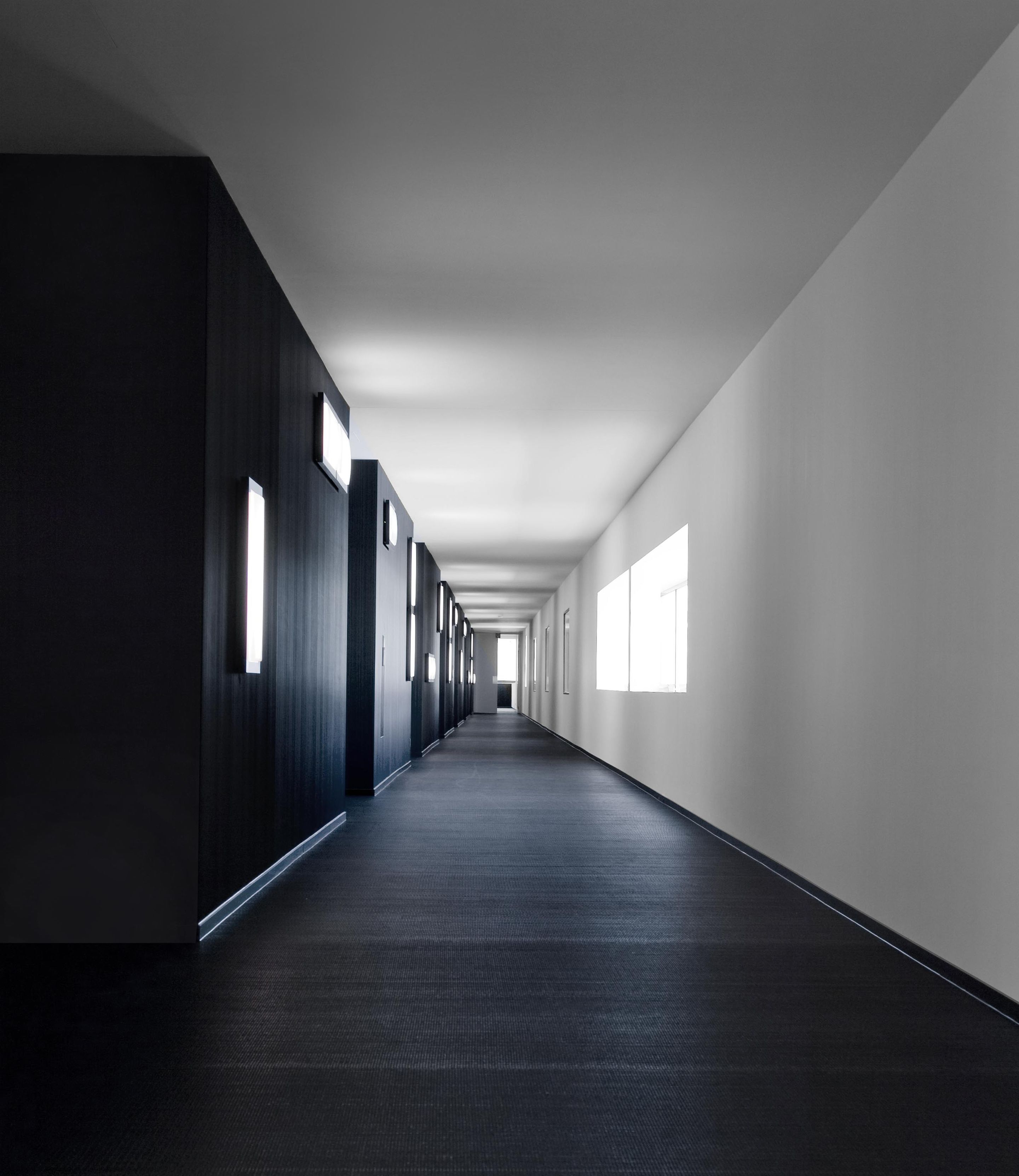 A long, narrow hallway with dark walls and a dark floor, leading to a bright exit at the end.
