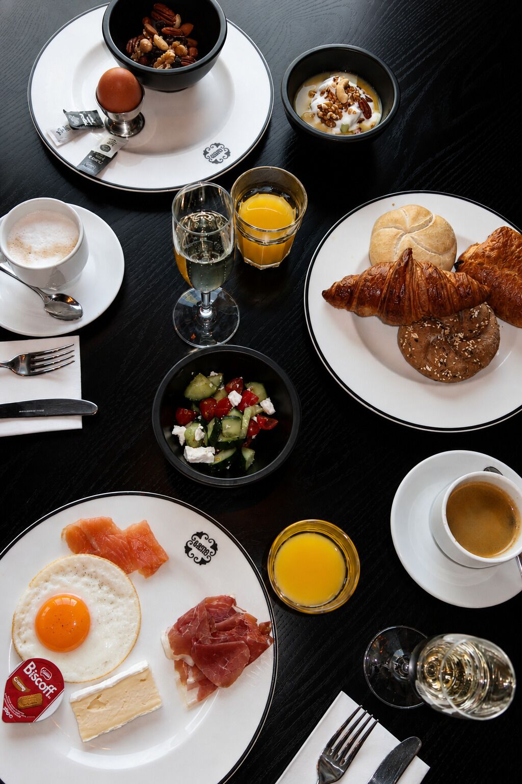 Una mesa de desayuno con platos de comida, incluyendo huevos, salmón ahumado y cruasanes, junto con bebidas como café y jugo de naranja.
