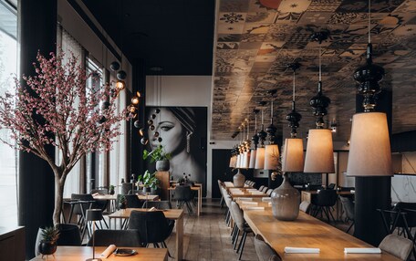 A modern restaurant with long wooden tables, black chairs, and hanging pendant lights.