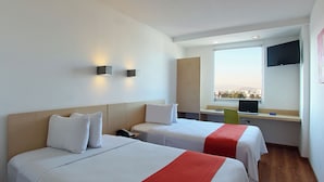 Habitación de hotel con dos camas, un escritorio con una computadora portátil, un televisor y una ventana con vista a la ciudad.