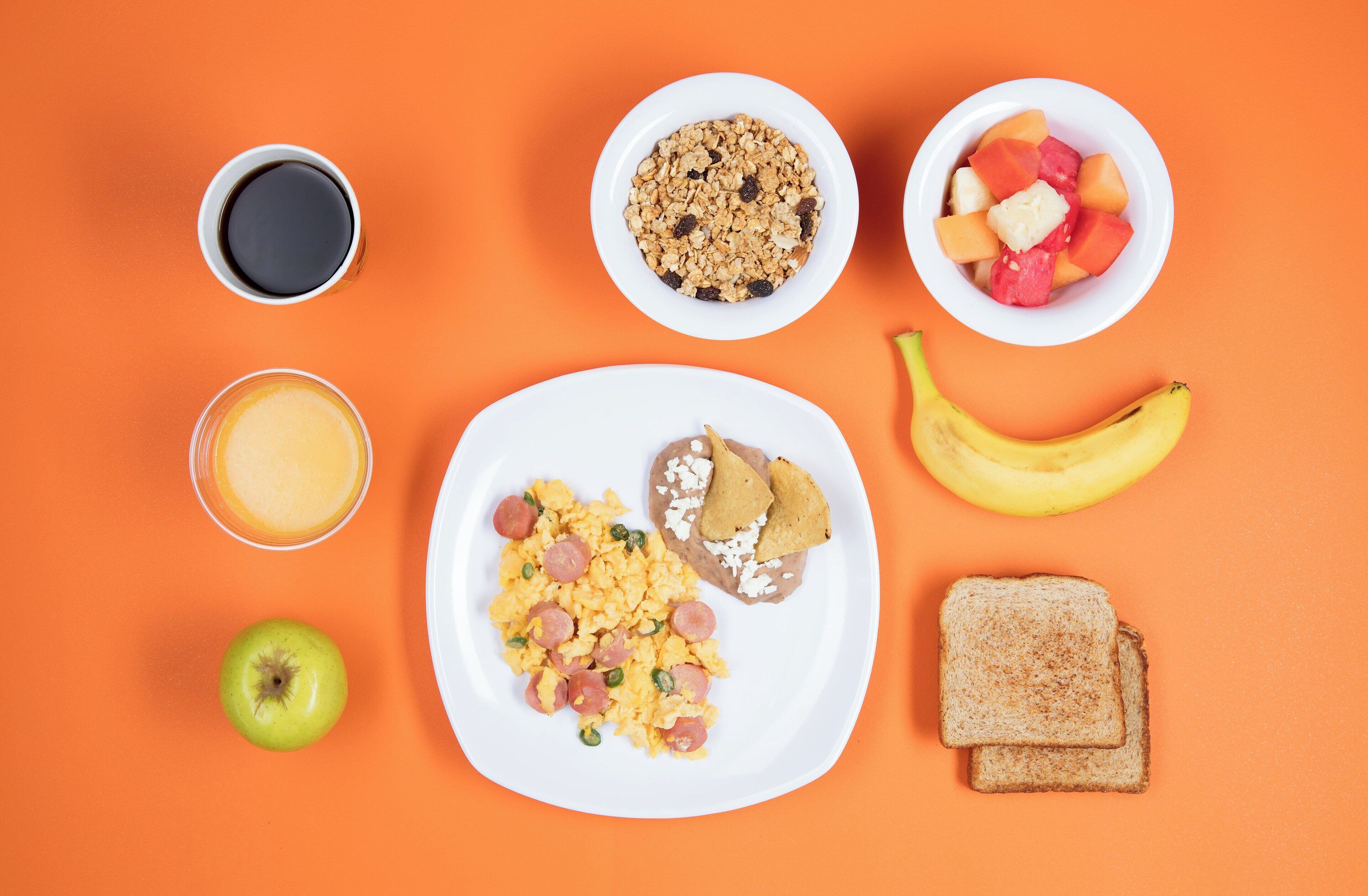 A breakfast plate with scrambled eggs, sausage, and toast, a banana, a green apple, a glass of orange juice, a bowl of granola, a bowl of fruit, and a cup of coffee on an orange background.