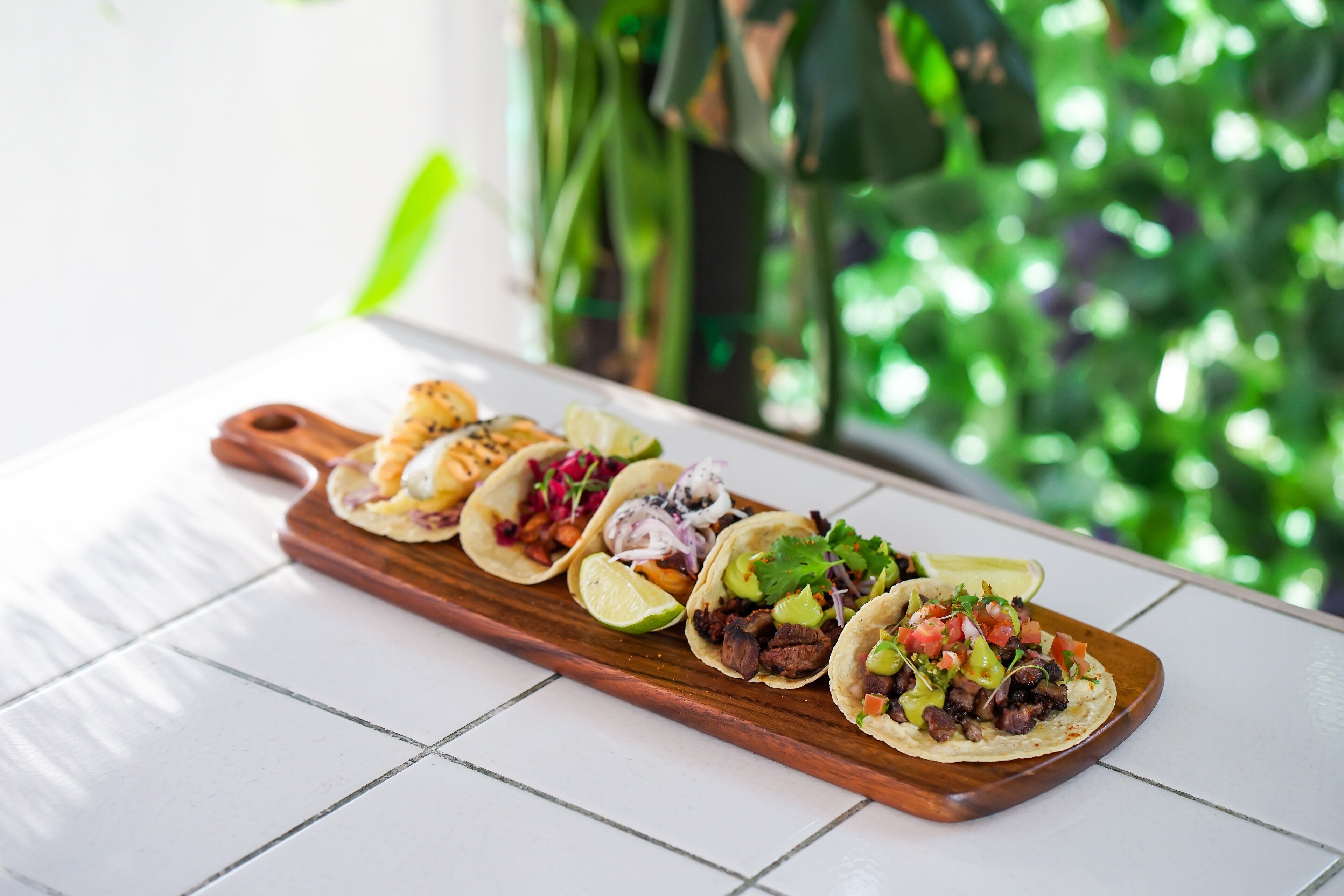 Une planche de service en bois sur laquelle sont disposés trois tacos, le tout posé sur une surface carrelée.