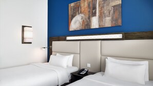 Habitación de hotel con dos camas, una mesita de noche con teléfono, una lámpara de pared y un cuadro abstracto grande.