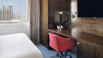 A hotel room with a bed, a desk with a chair, a TV, and a city view through the window.