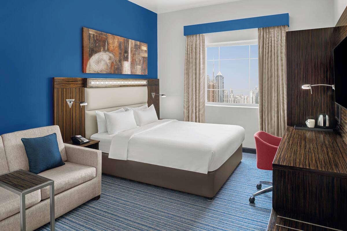 Habitación de hotel con una cama grande, un escritorio, una silla, un televisor y vistas a la ciudad.