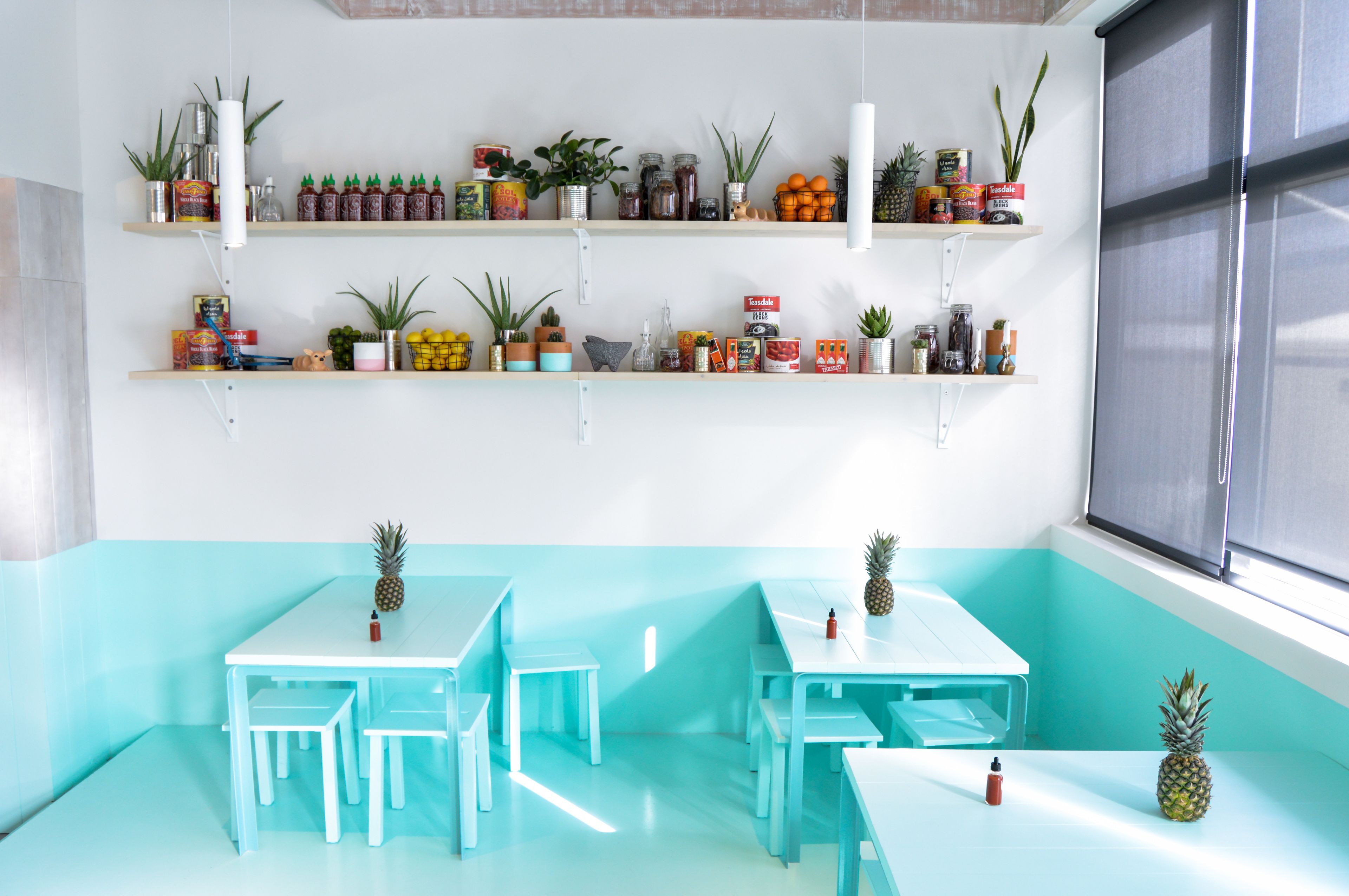 A room with teal walls and white furniture, featuring two tables and chairs, a window with blinds, and shelves with various items.