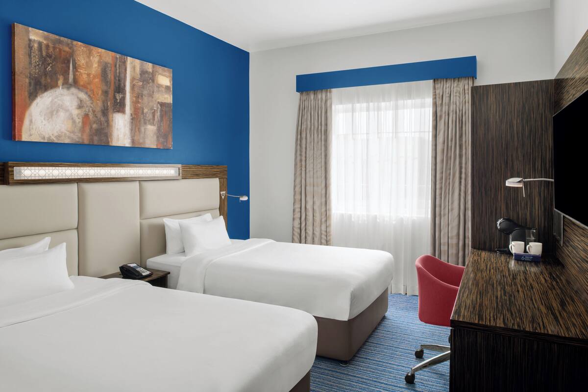 Habitación de hotel con dos camas, un escritorio, una silla roja, un cuadro en la pared y un ventanal con cortinas.