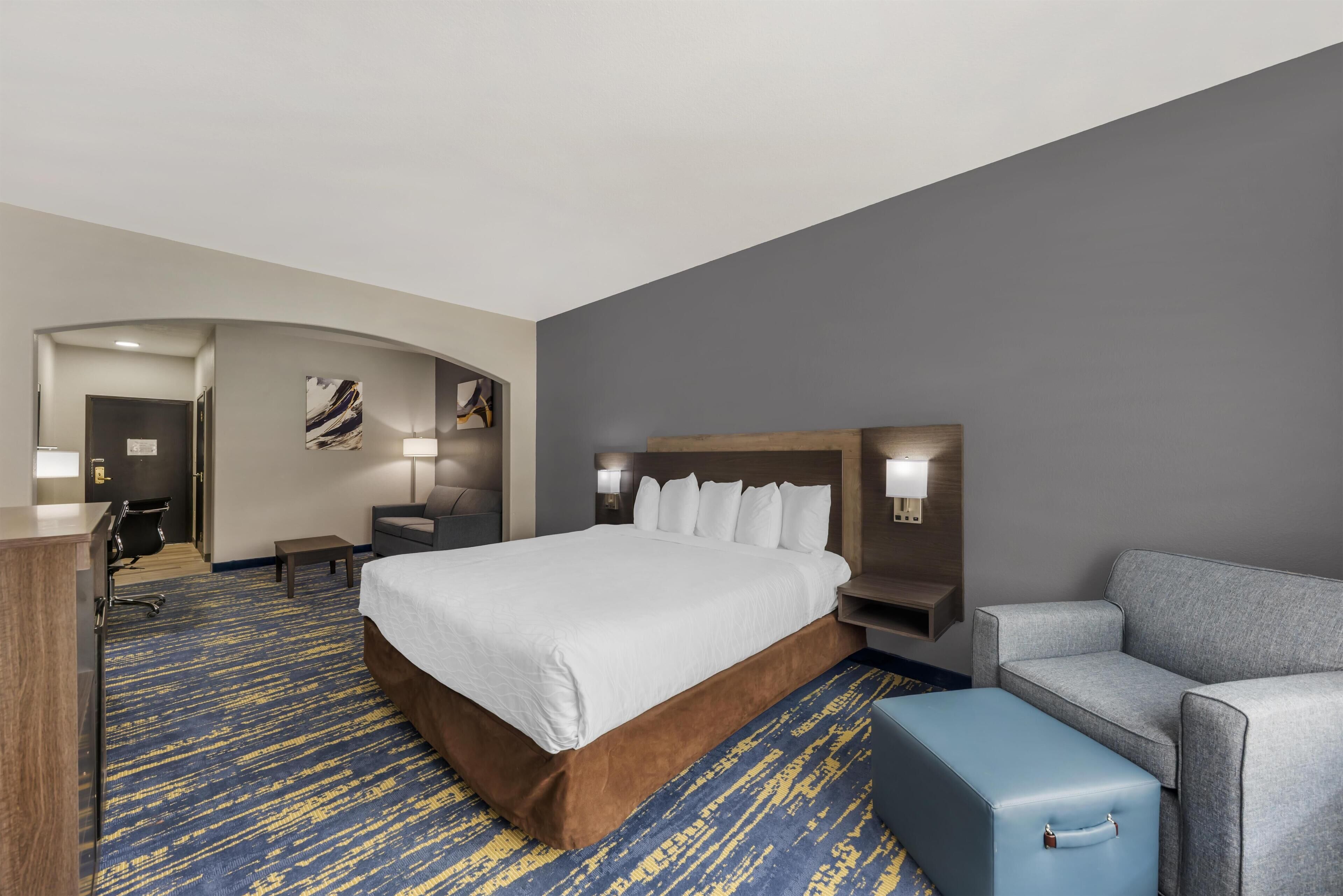 A hotel room with a large bed, a grey sofa, a blue suitcase, and a desk with a lamp.