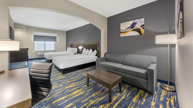 Habitación de hotel con techo curvo, dos camas, sofá gris, mesa de centro de madera y alfombra azul y amarilla estampada.