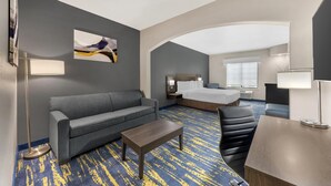 Habitación de hotel con sofá gris, una pequeña mesa de madera, un escritorio, cama con cabecera blanca y un ventanal grande.