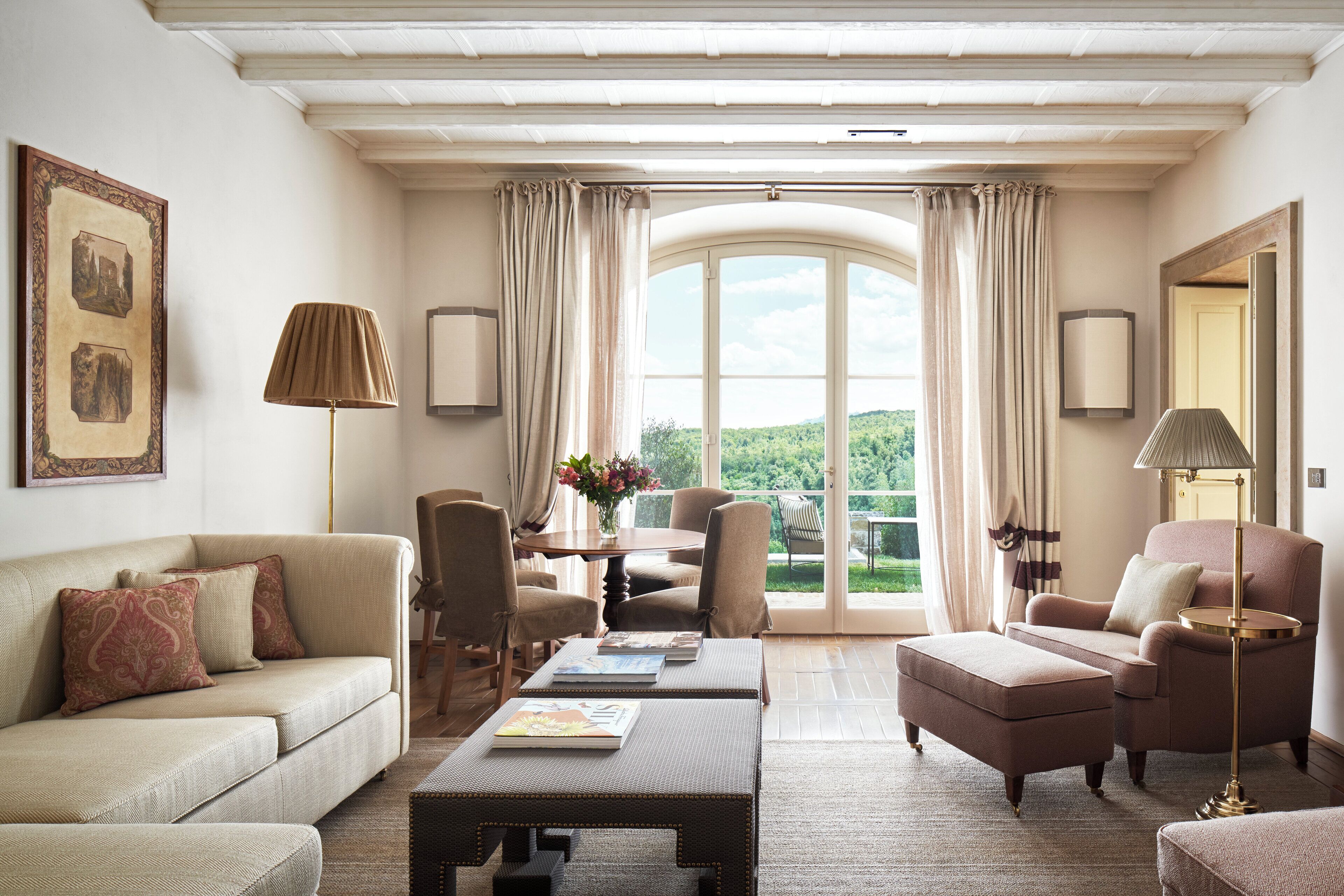 A spacious living room with a sofa, armchairs, a coffee table, and a dining area with a table and chairs. A large window with curtains provides a view of the outdoors.
