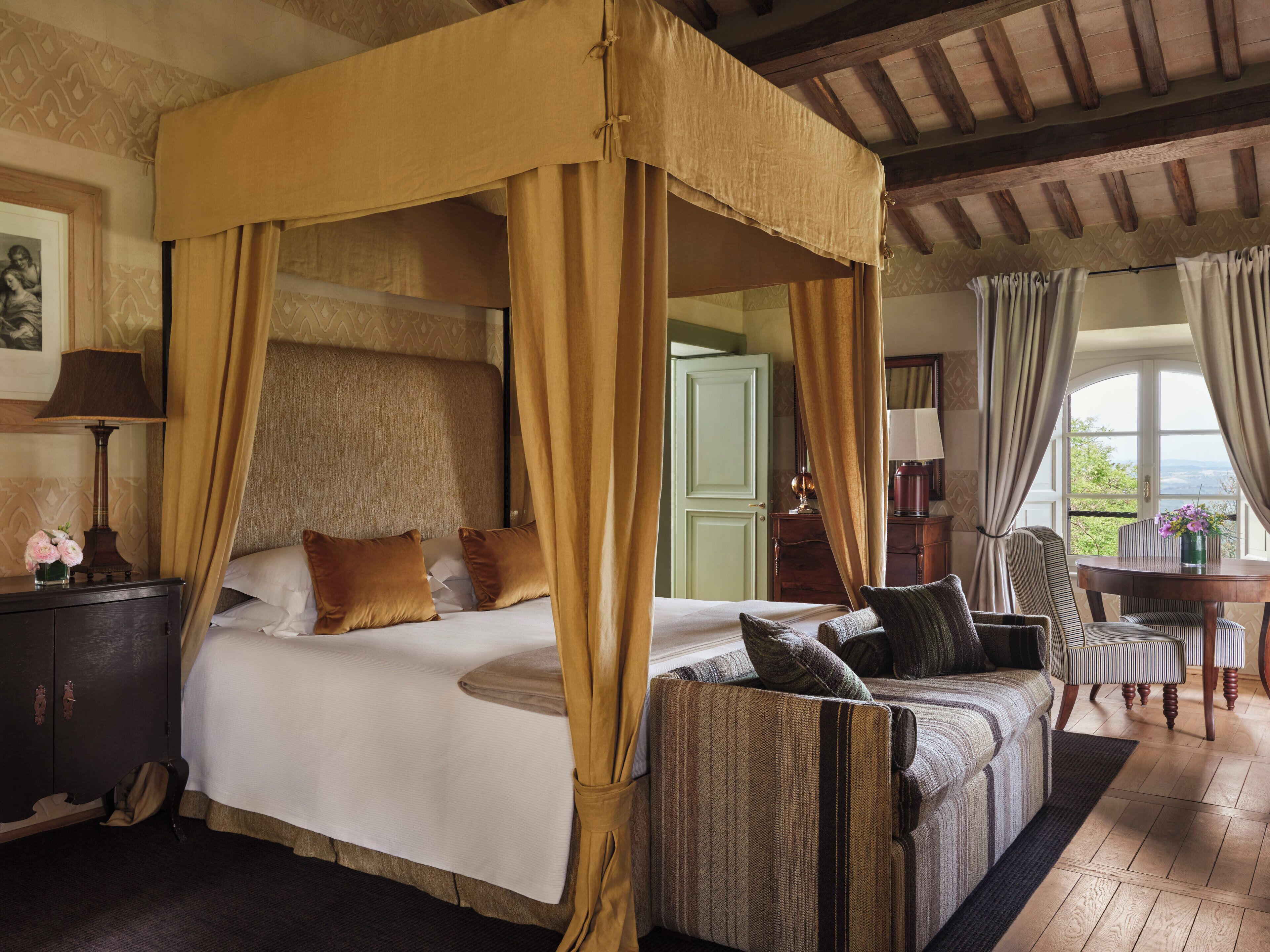 A four-poster bed with a canopy, a sitting area with a chair and table, and a view through a window.