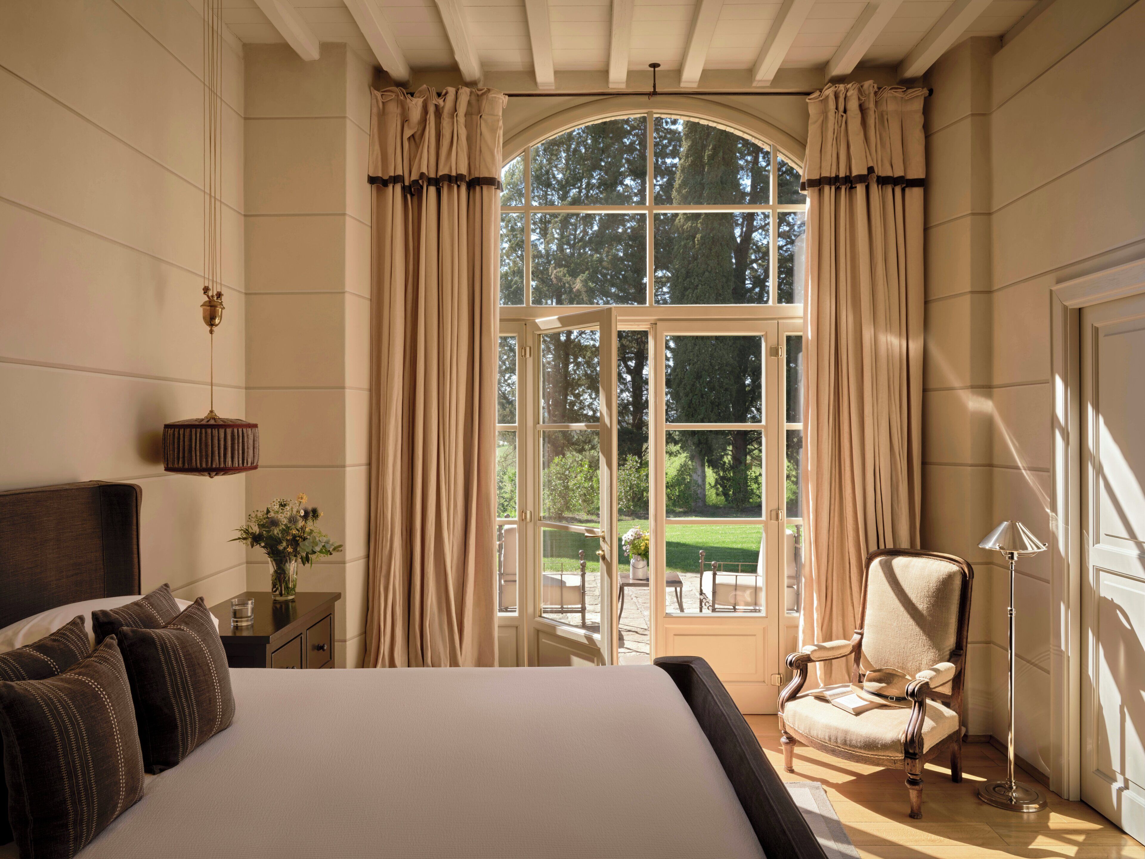 Ein Schlafzimmer mit einem großen Bett, einem Sessel, einem Nachttisch, einer Lampe und einem Fenster mit Vorhängen.