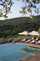 Una piscina infinita con sillones de descanso y sombrillas, rodeada por una ladera boscosa.