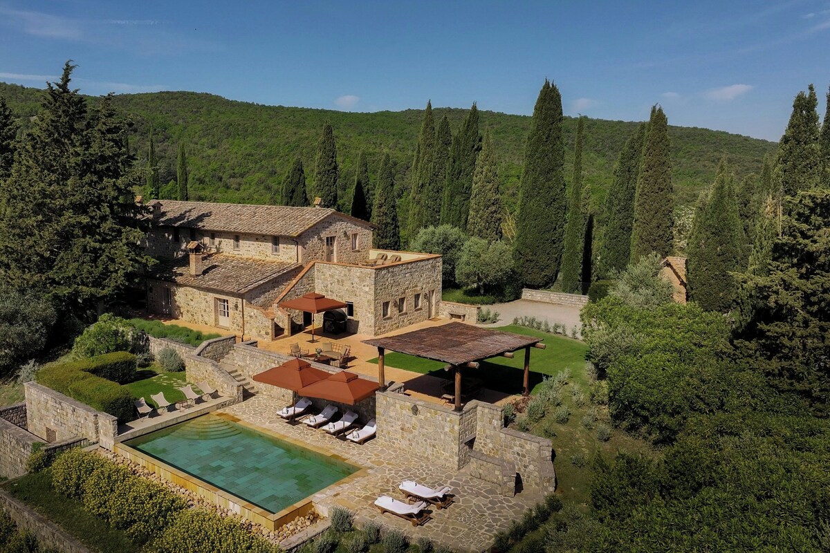 Una villa de piedra con piscina, mobiliario exterior y vistas a una ladera boscosa.
