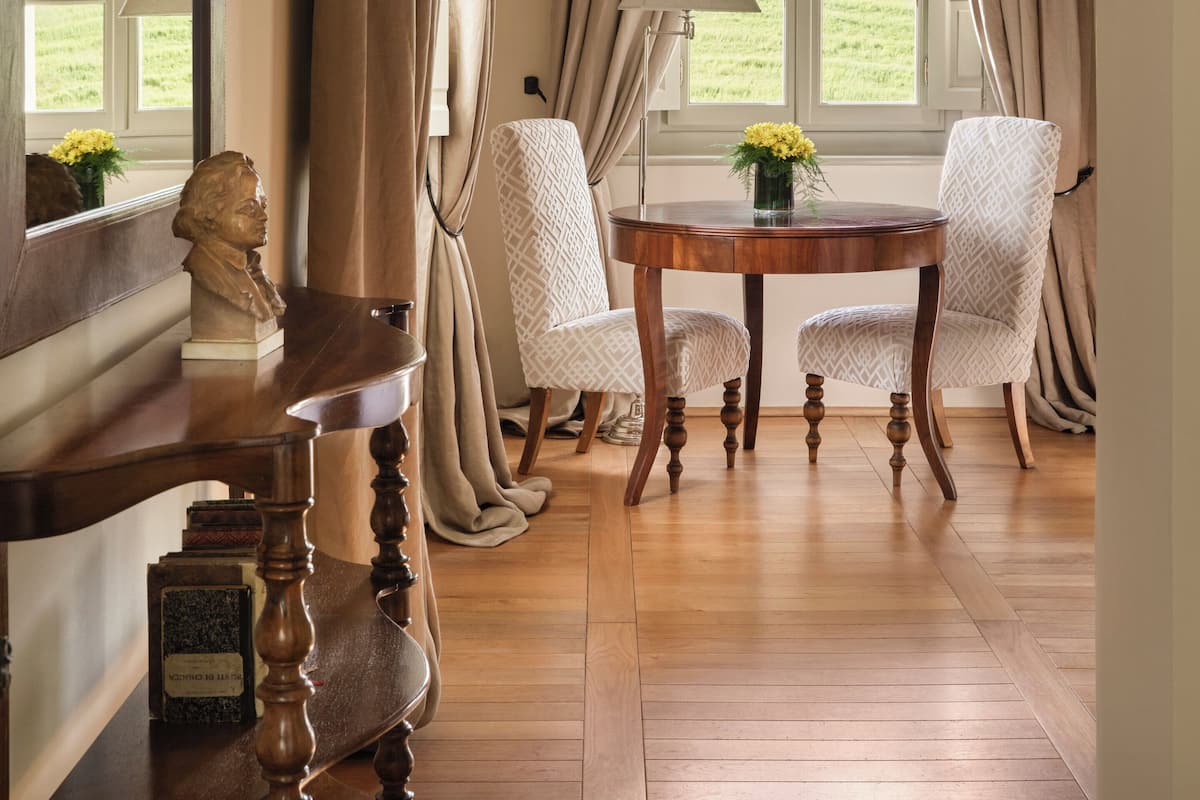 Un comedor con una mesa de madera, dos sillas estampadas, una lámpara y un busto sobre la mesa.