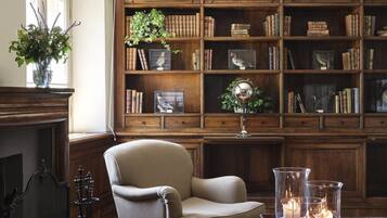 A library with wooden bookshelves, a fireplace, a comfortable armchair, and a vase with flowers.