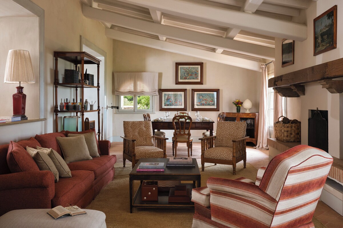 A cozy living room with a red sofa, a striped armchair, and a coffee table with books. There is a bookshelf, a fireplace, and framed artwork on the wall.