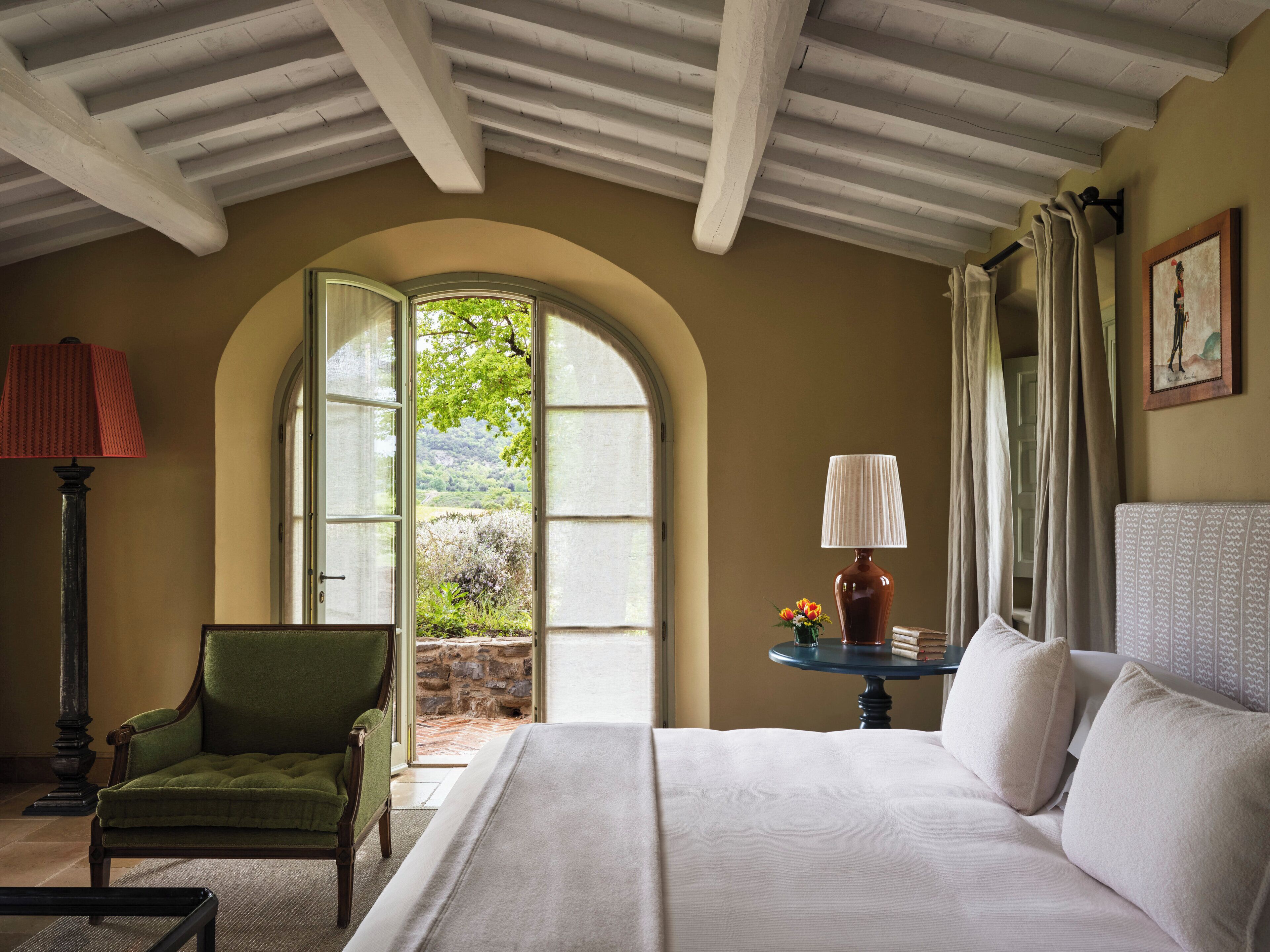 Un dormitorio con una cama grande, un sillón verde, una mesa con lámpara y vistas al verdor a través de una ventana abierta.