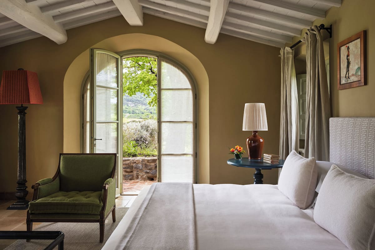 Un dormitorio con una cama grande, un sillón verde, una mesa con lámpara y vistas al verdor a través de una ventana abierta.