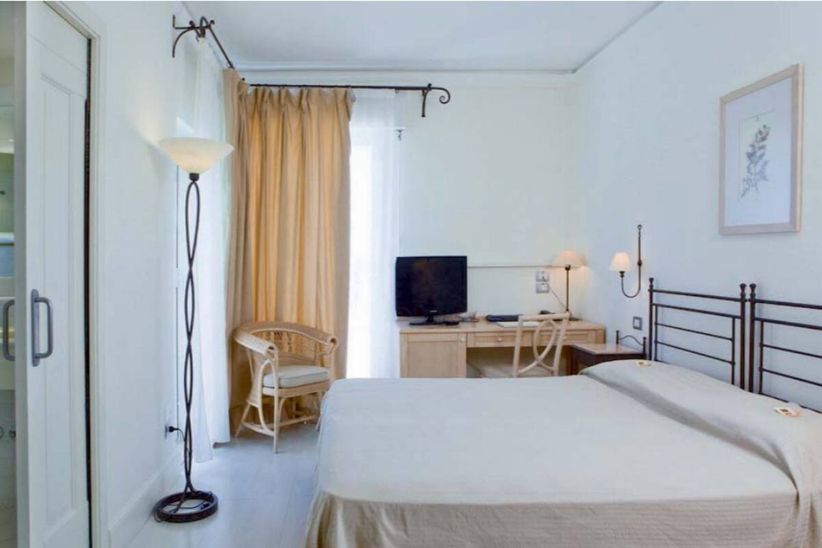 A hotel room with a bed, a desk with a computer, a chair, a television, and a floor lamp.
