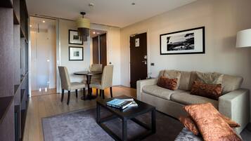 Una sala de estar moderna con sofá, mesa de centro y sillones. Hay un comedor con mesa y sillas. Un cuadro cuelga de la pared y una lámpara está sobre una mesita auxiliar.