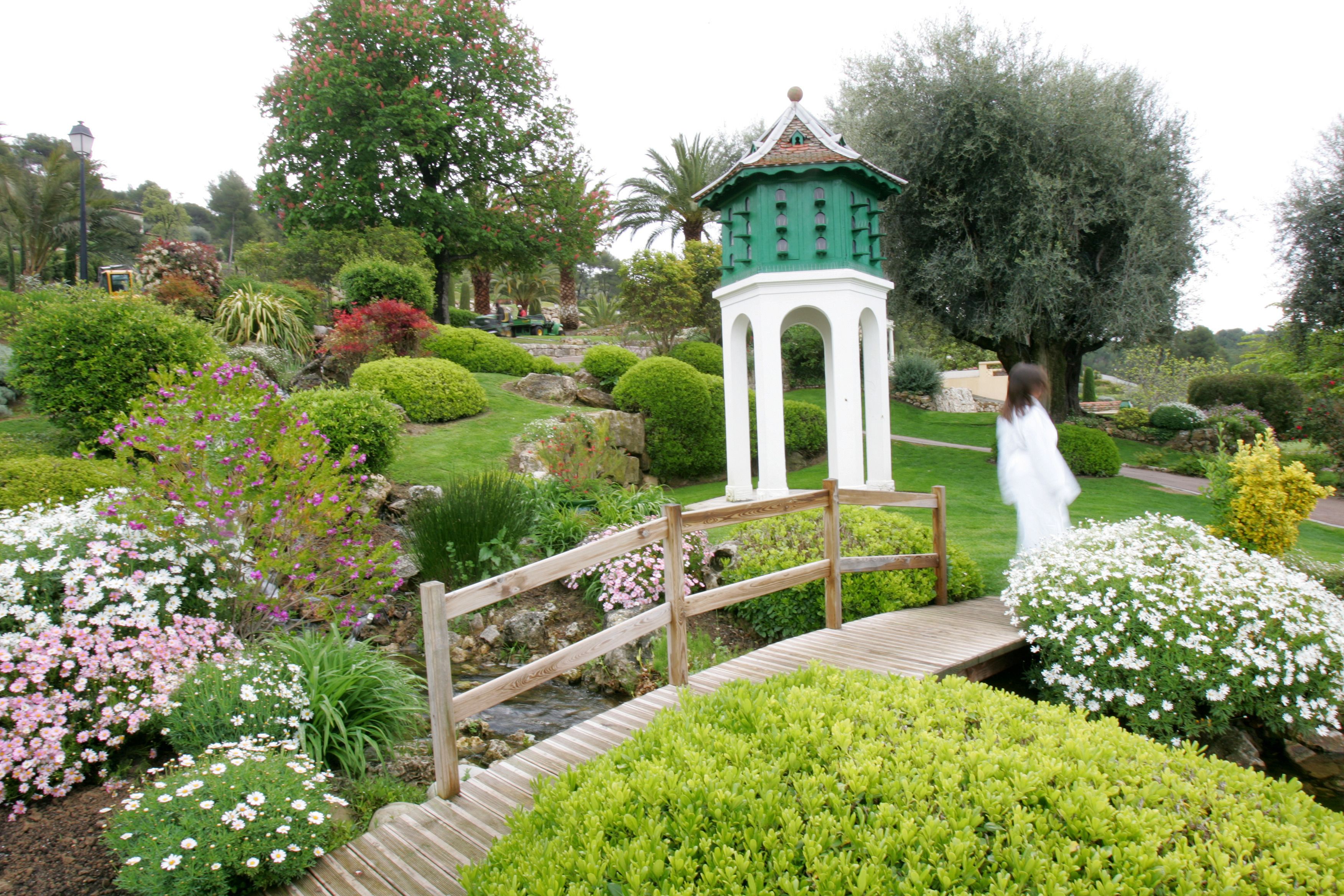 A garden with a wooden bridge, a white gazebo, and various flowering plants.