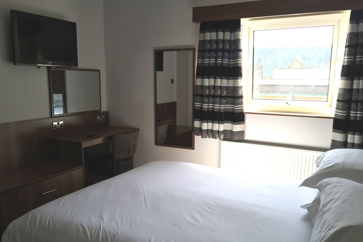 A hotel room with a bed, a desk, a mirror, a TV, and a window with curtains.