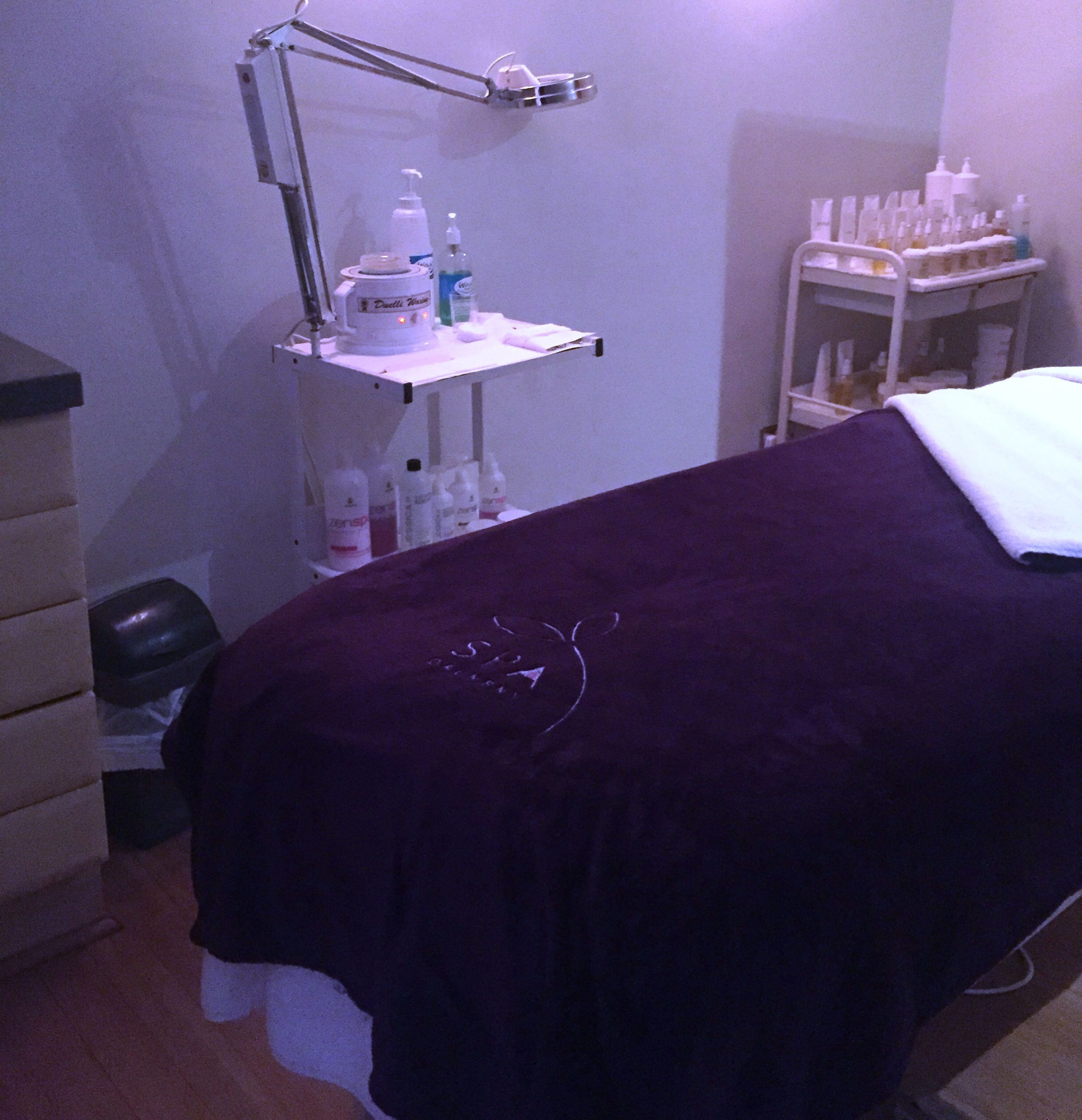 A room with a bed covered in a dark purple blanket, a white bedside table with a lamp, and a shelf with various toiletries.