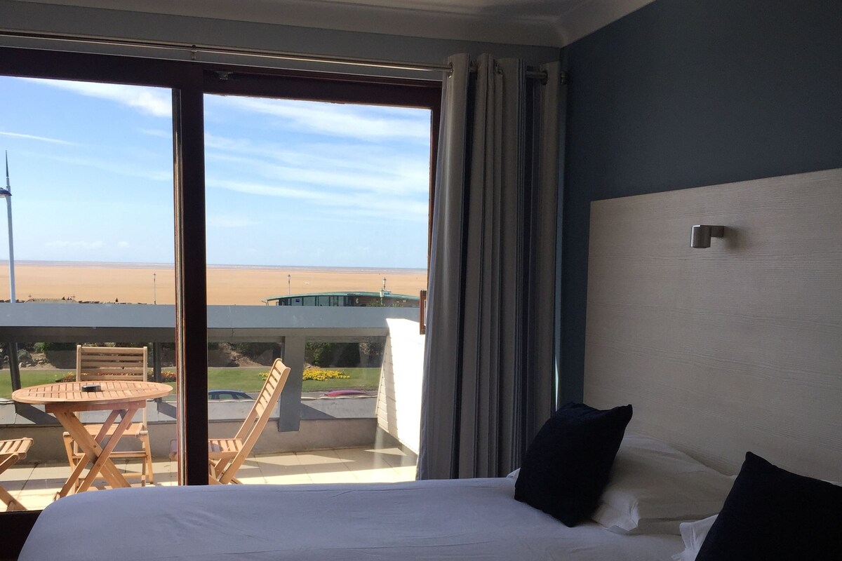 A hotel room with a large bed, a view of the beach, and a balcony with a table and chairs.