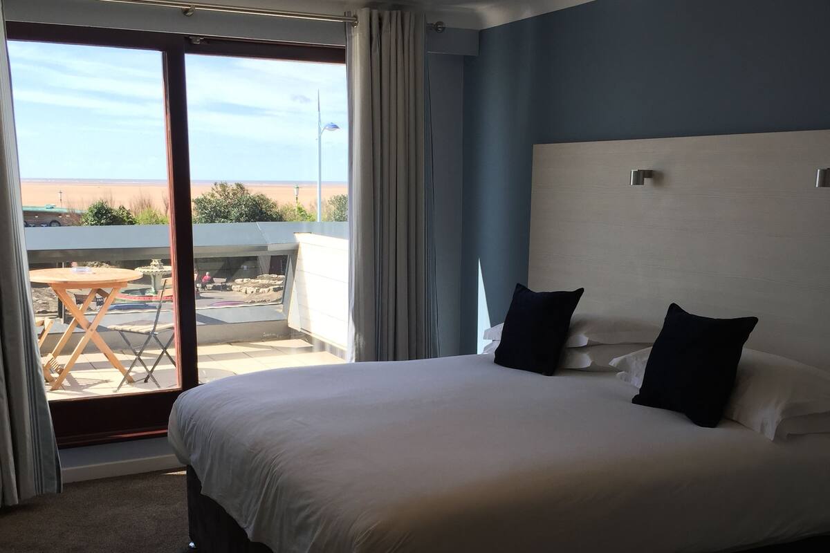 A hotel room with a large bed, a view of the beach, and a balcony with outdoor furniture.