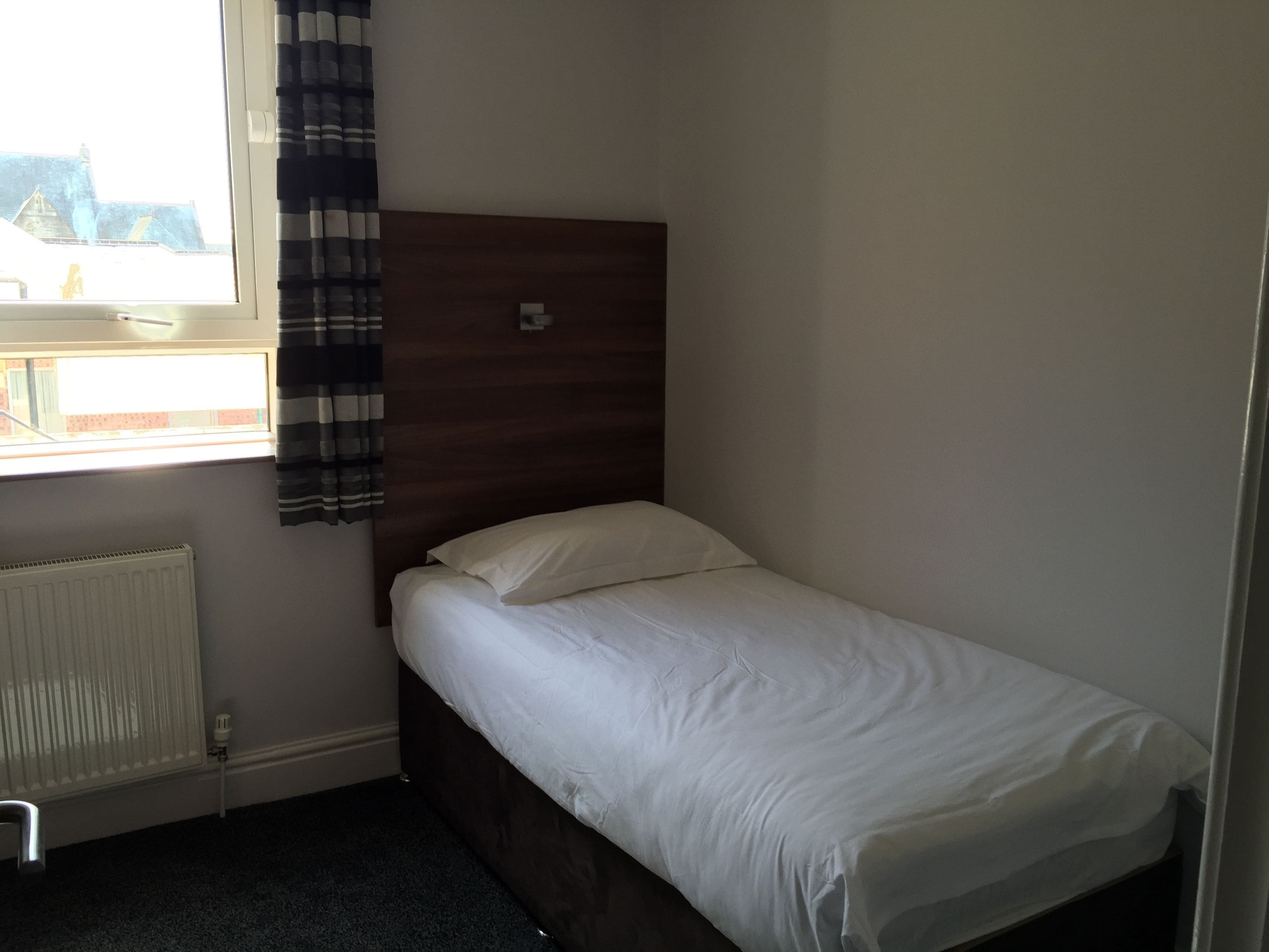 A small, single-bed room with a wooden headboard, a window with blinds, and a radiator.