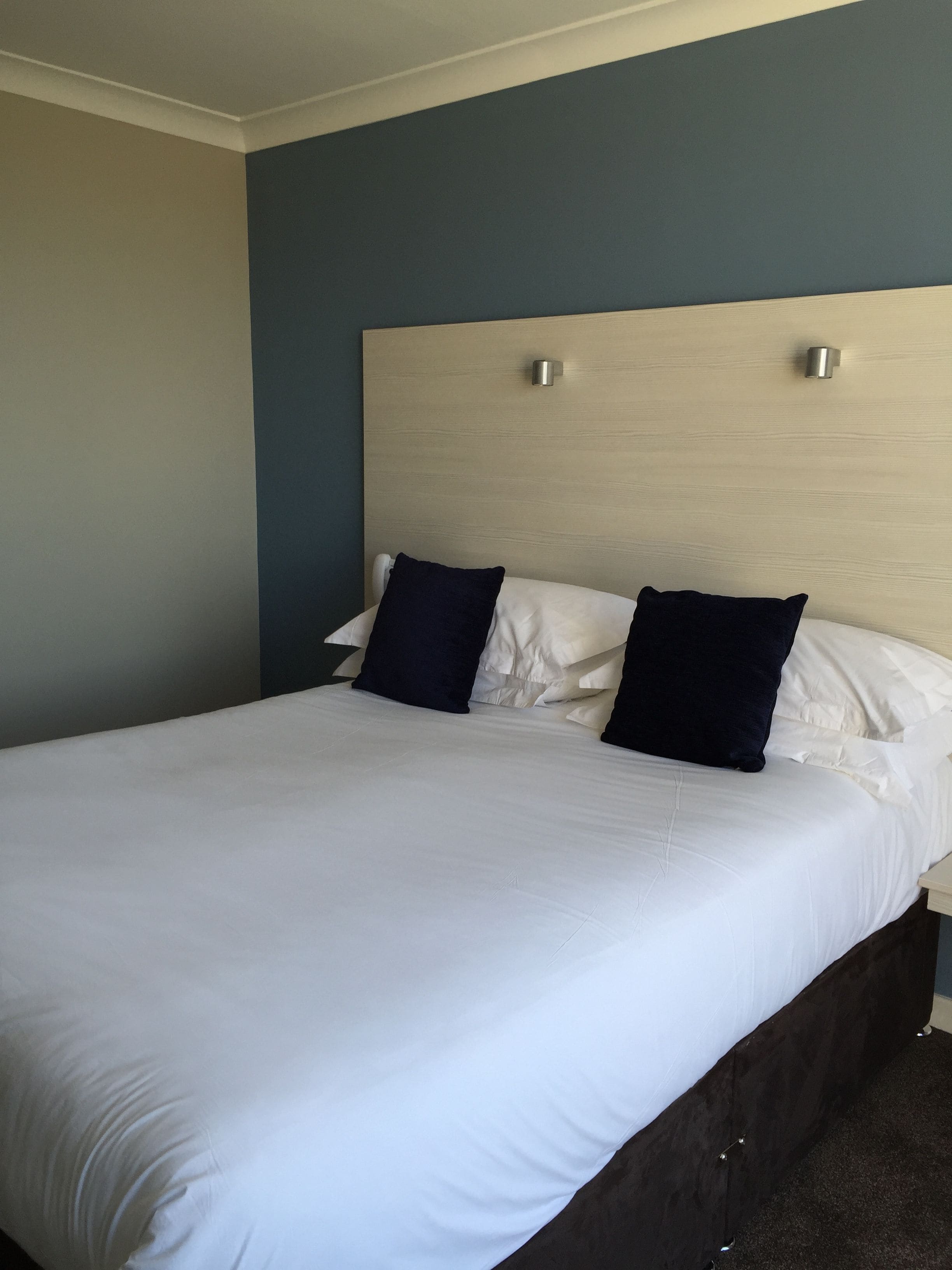 Een groot bed met witte lakens en twee donkere kussens, een houten hoofdbord en een eenvoudige muur.
