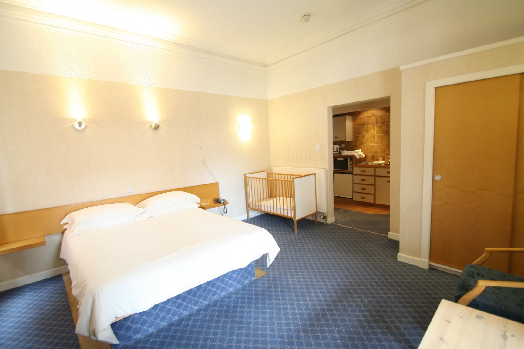 A hotel room with a large bed, a crib, a desk, and a kitchenette.