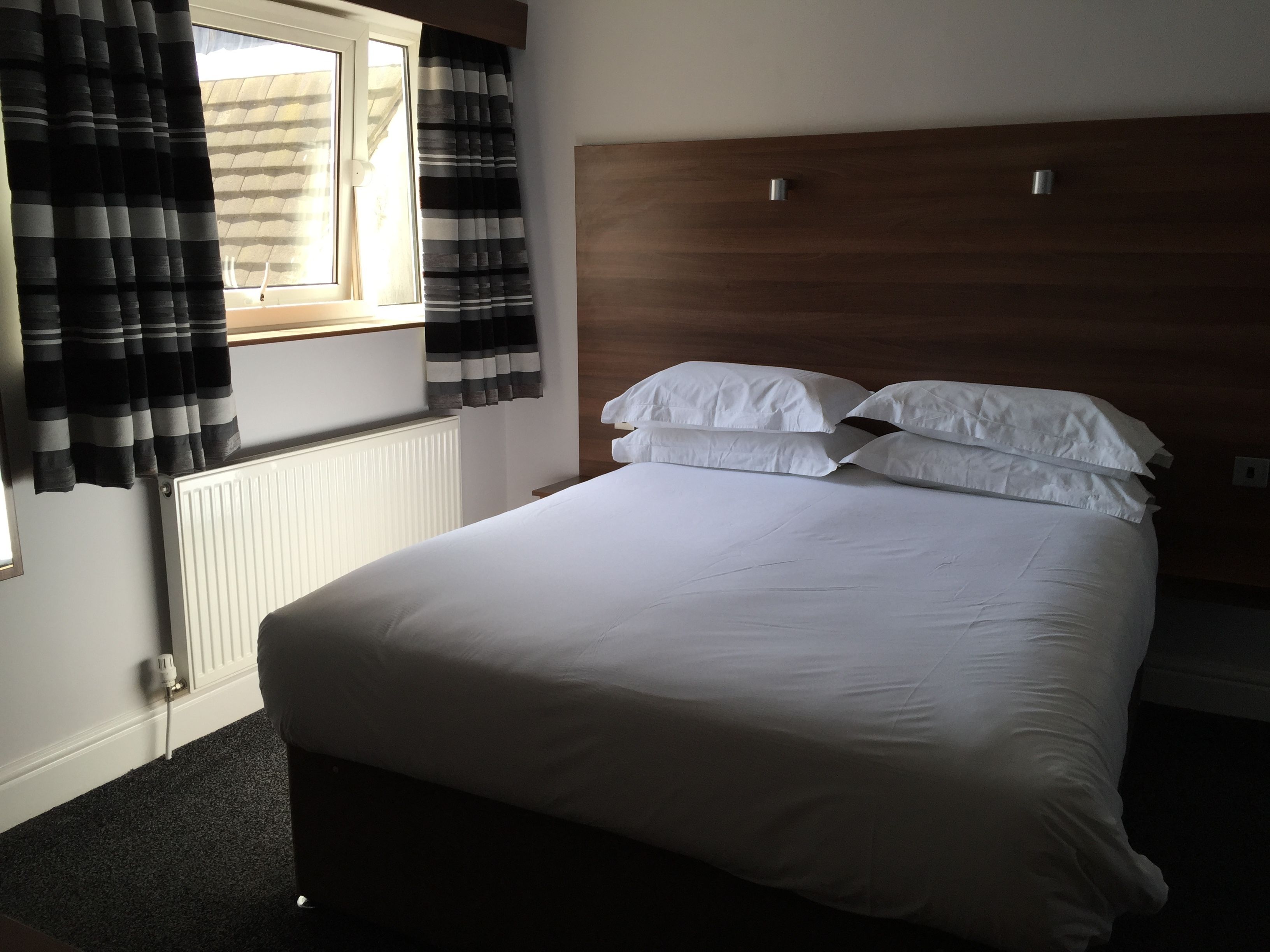 A bedroom with a wooden headboard, white bedding, a window with curtains, and a radiator.