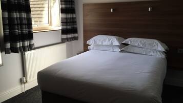 A bedroom with a wooden headboard, white bedding, a window with curtains, and a radiator.