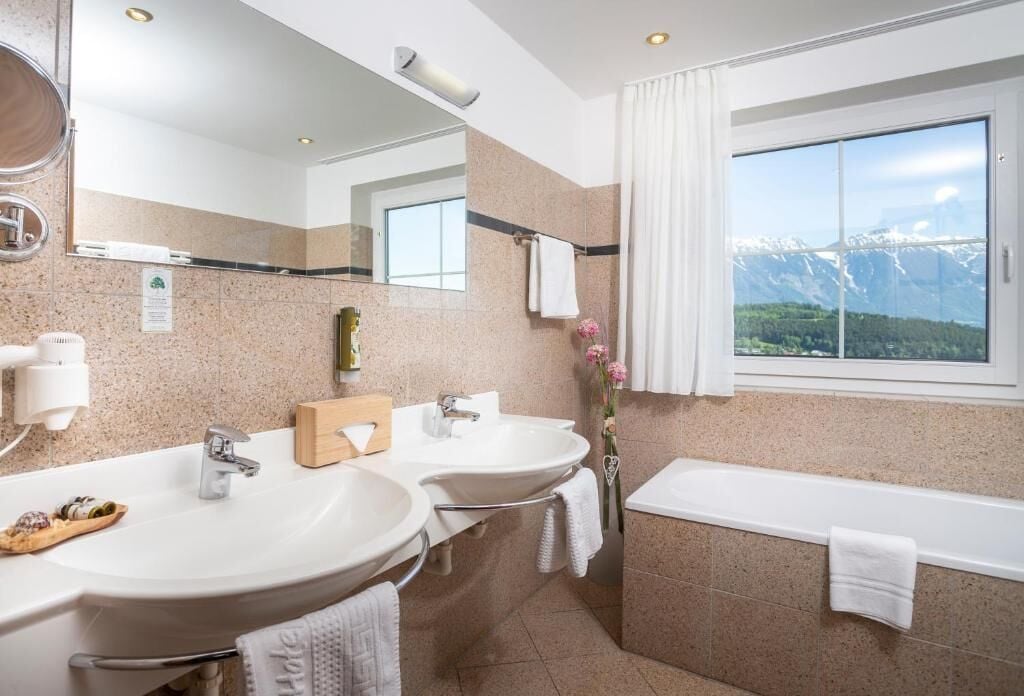 A modern bathroom with two sinks, a bathtub, a large mirror, and a window with a mountain view.