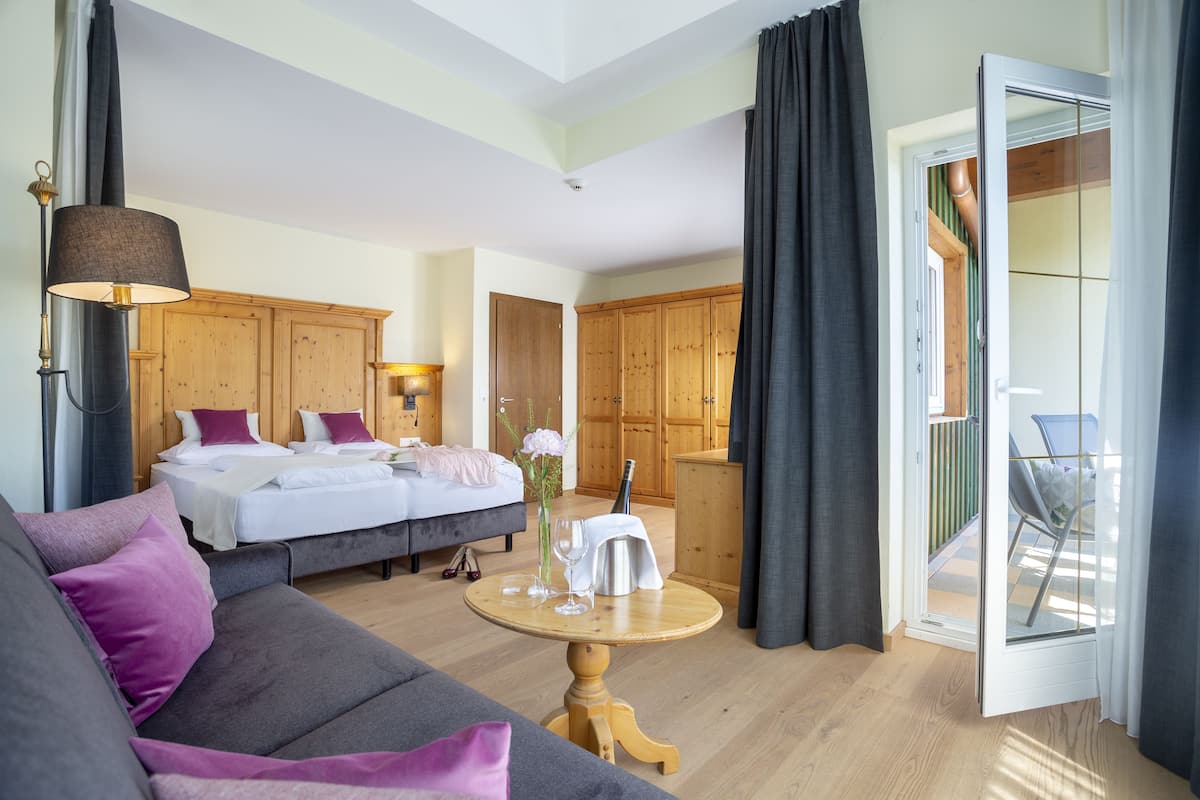 A hotel room with a bed, a sofa, a table with a bottle and glasses, wooden cabinets, and a balcony with a chair.