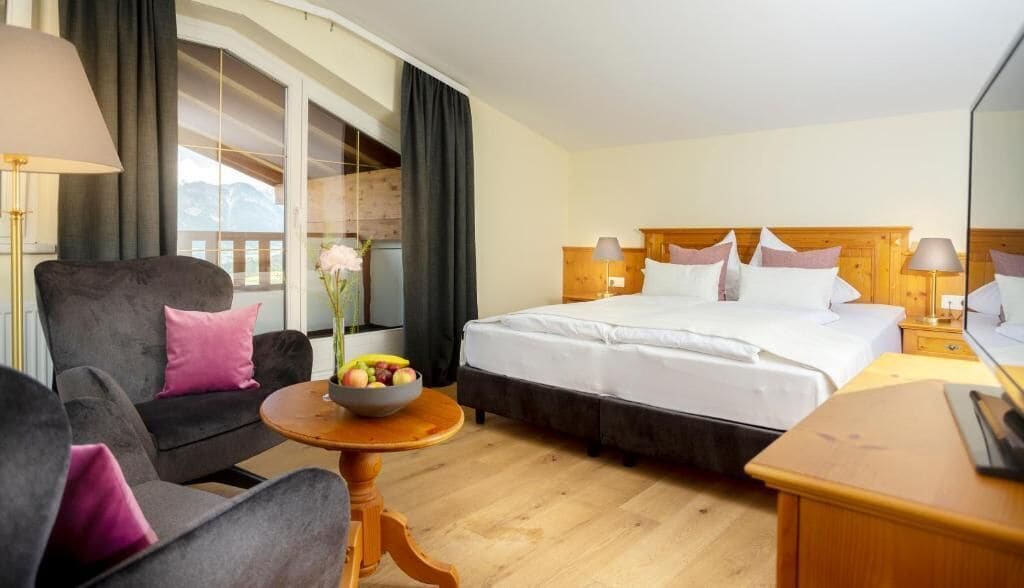 Une chambre d’hôtel avec un lit, un canapé, une petite table avec des fruits, et une vue sur les montagnes par une grande fenêtre.