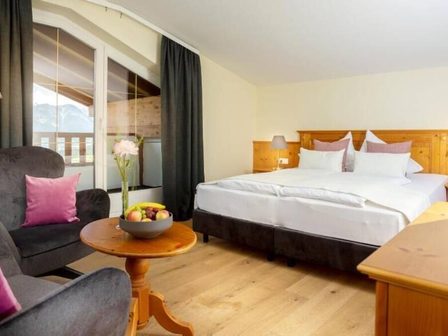 Habitación de hotel con cama, sofá, una mesita con fruta y vistas a montañas a través de un amplio ventanal.