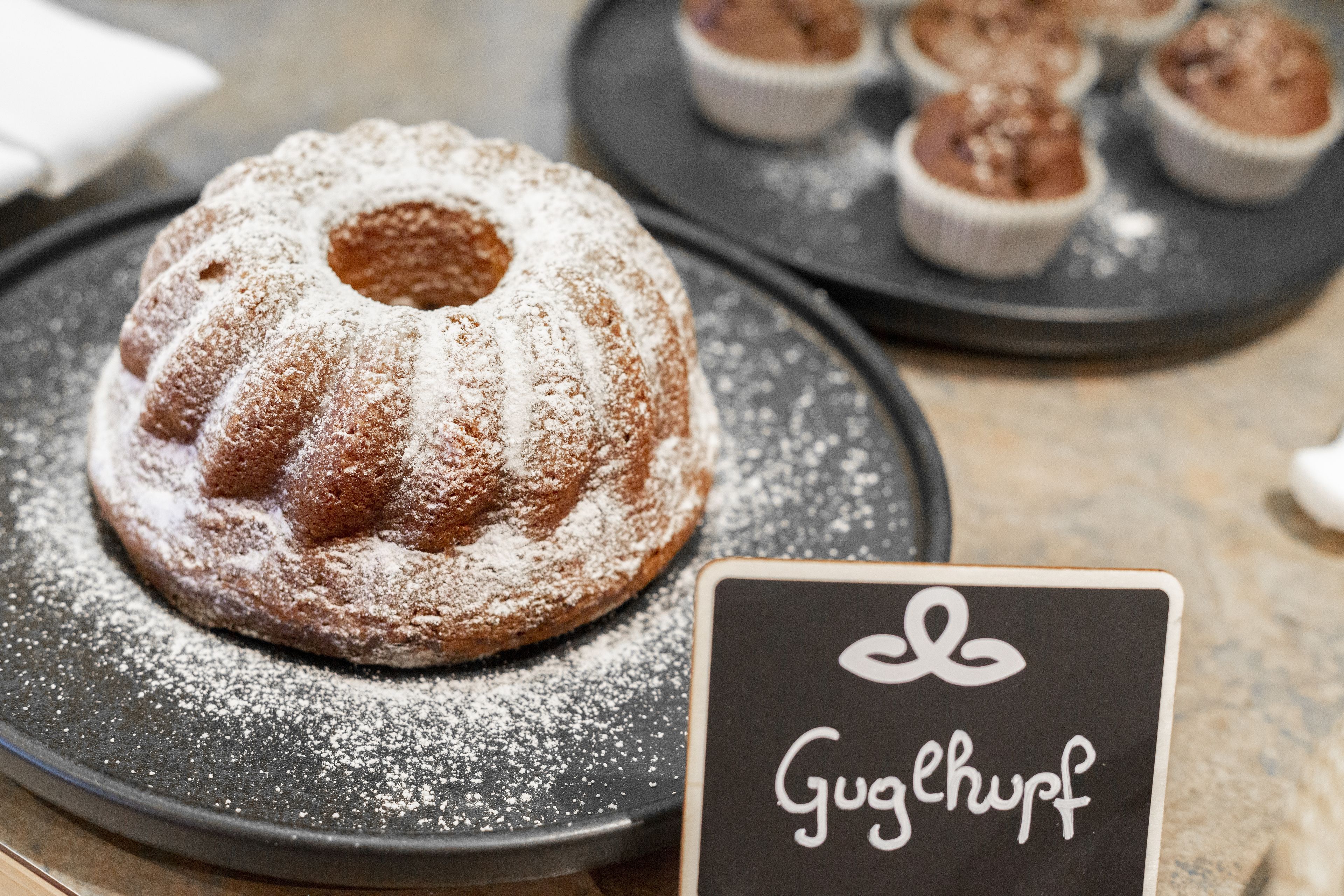 A bundt cake with powdered sugar on a plate, a sign with 'Gugelhupf' written on it, and cupcakes in the background.